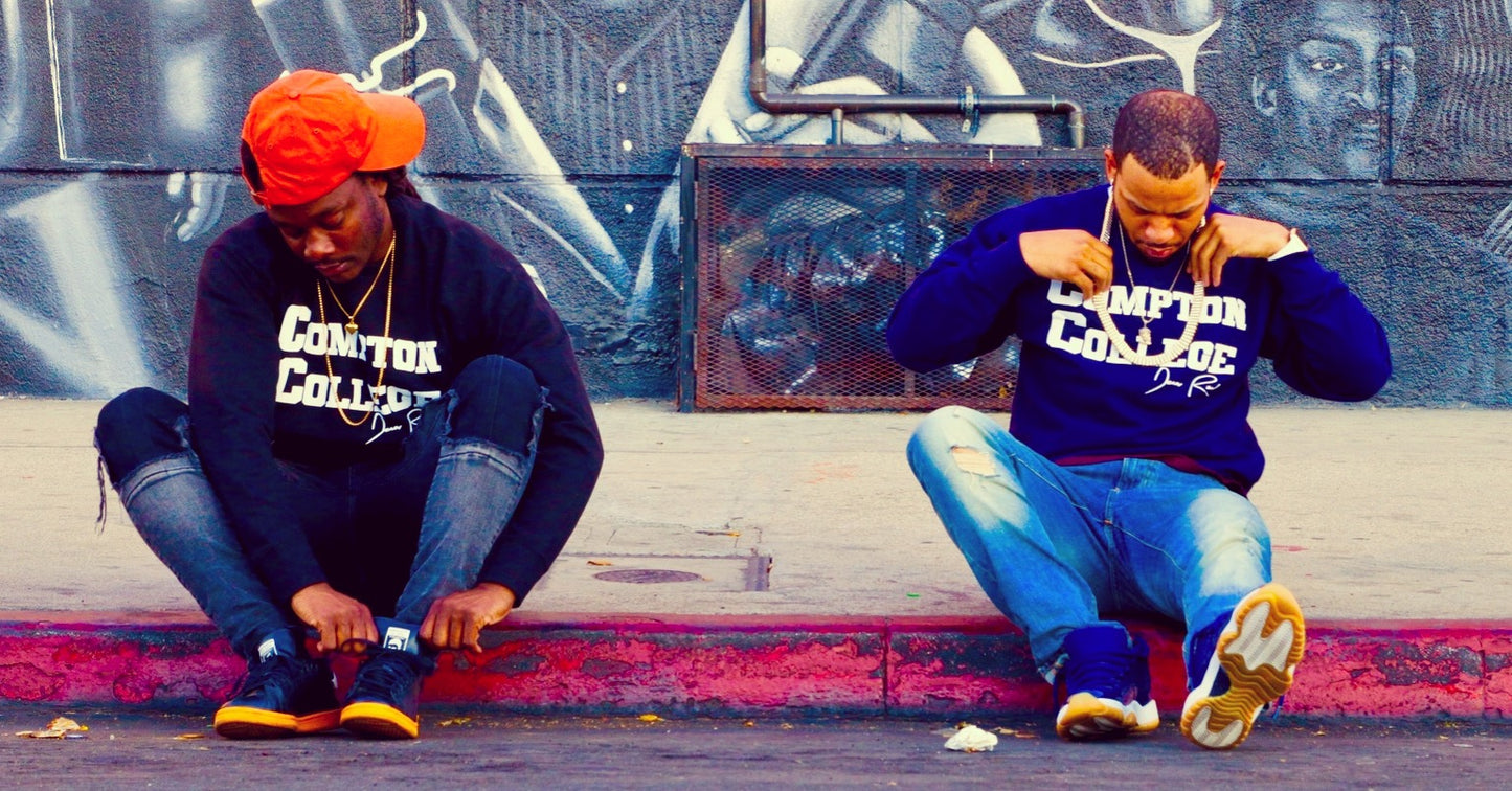 Two people sitting on a ledge wearing matching sweatshirts with 'Compton College' printed on them, in front of a graffiti-covered wall.