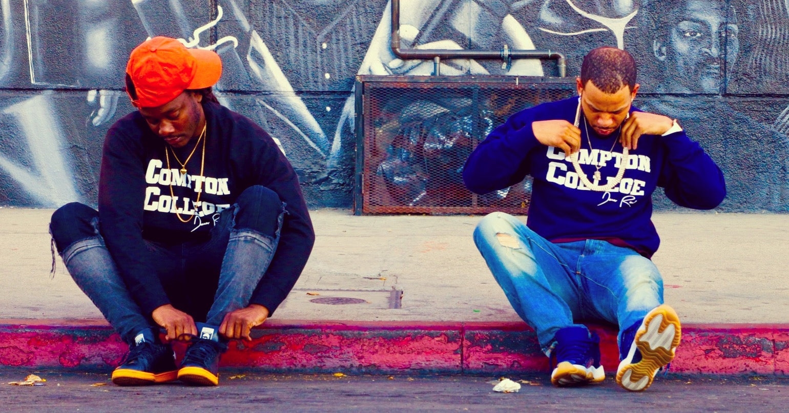 Two people sitting on a ledge wearing matching sweatshirts with 'Compton College' printed on them, in front of a graffiti-covered wall.